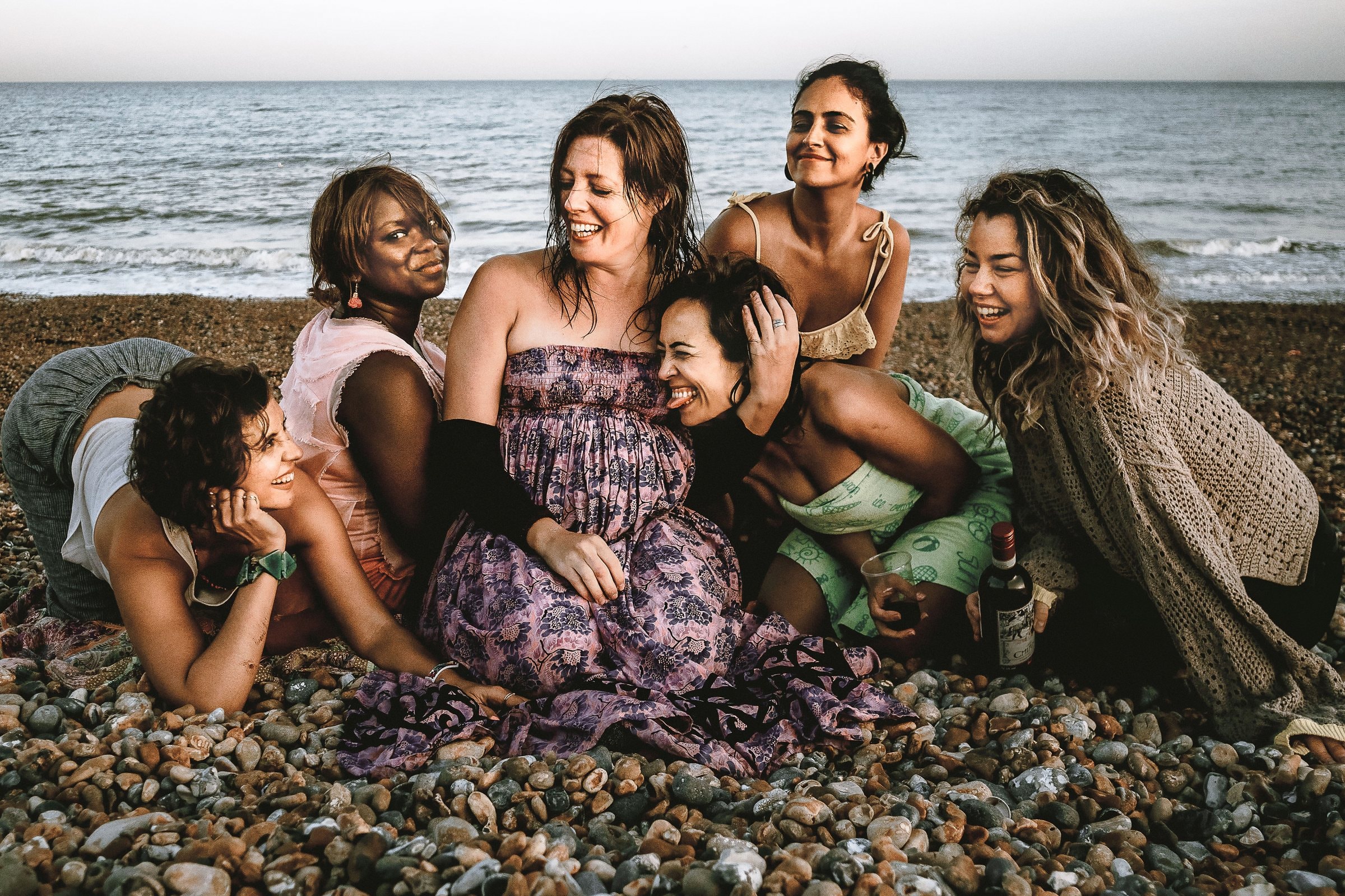 A group of women gathered around a pregnant friend on a pebble beach